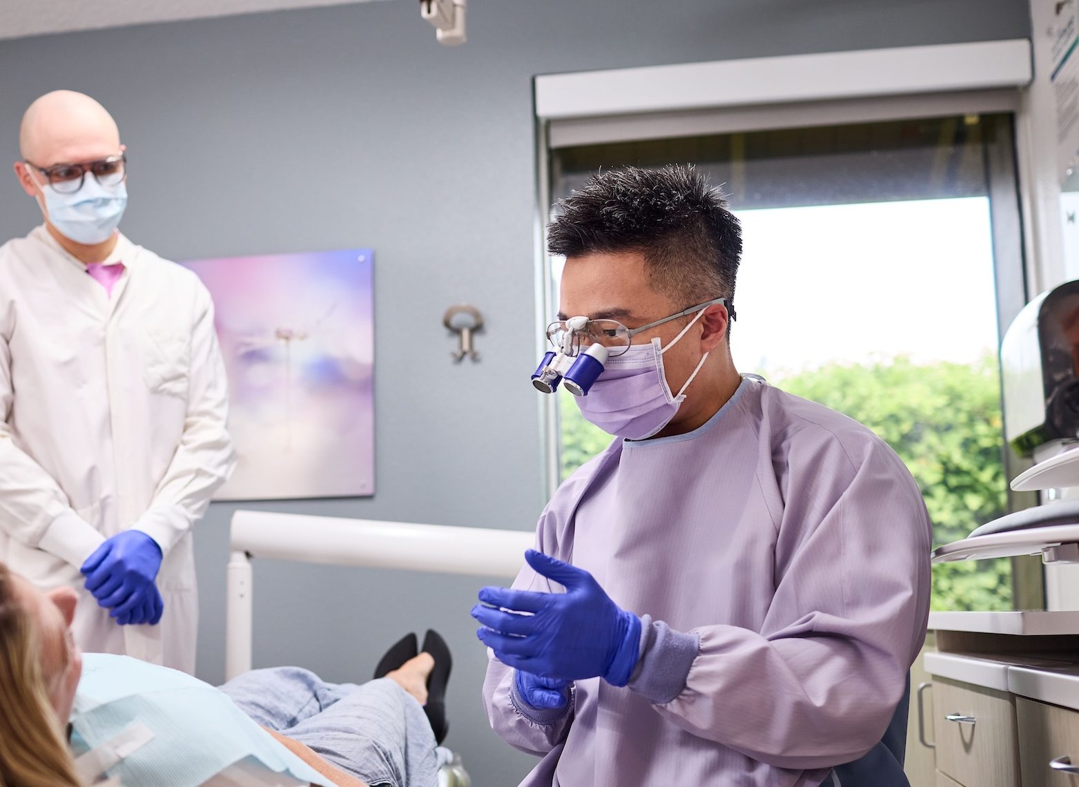 A dentist wearing dental loupes explaining something to a patient while another doctor observes